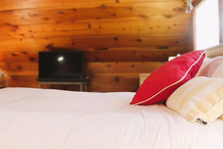 Bed-level perspective looking across white linens toward a flat-screen television in a wood-paneled room.