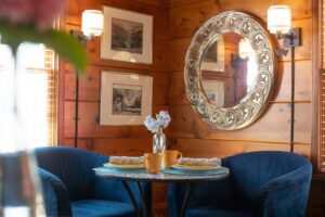 A cozy breakfast table for two flanked by blue velvet armchairs in a rustic pine-walled dining room.