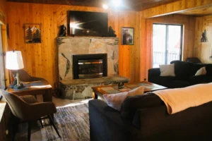 Cozy living room at Aspen Escape in Shaver Lake with a large stone fireplace, flat-screen TV, dark sofas, and wood paneling.